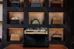 A luxurious black Hermès Birkin bag with palladium hardware and made with Horse Hair Troika displayed on a leather surface, set against a dark marble wall featuring a backlit collection of various Hermès handbags in different colors and materials.