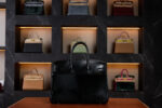 A luxurious black Hermès Birkin bag with palladium hardware and made with Horse Hair Troika displayed on a leather surface, set against a dark marble wall featuring a backlit collection of various Hermès handbags in different colors and materials.